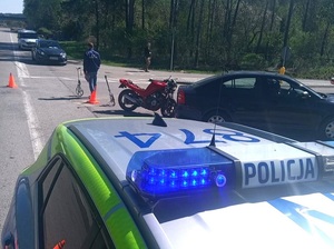 miejsce zdarzenia drogowego. Na jezdni przy skrzyżowaniu za granatową Skodą Octavią stoi czerwony motocykl. Obok stoi technik kryminalistyki, przy którym znajduje się przyrząd do pomiaru odległości. Na poboczu stoją dwa pojazdy osobowe. Na pierwszym planie, przed Skodą i motocyklem stoi oznakowany policyjny radiowóz