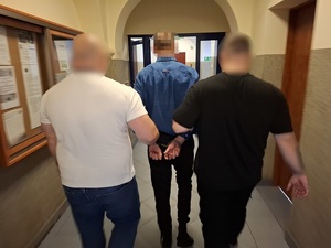 dwaj policjanci kryminalni prowadzą korytarzem mężczyznę, który ma ręce skute za plecami kajdankami