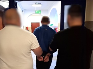 dwaj policjanci kryminalni prowadzą korytarzem mężczyznę, który ma ręce skute za plecami kajdankami