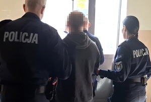 policjant i policjantka służby konwojowej wyprowadzają z budynku komendy zatrzymanego mężczyznę