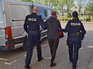 policjant i policjantka służby konwojowej prowadzą w kierunku nieoznakowanego radiowozu zatrzymanego mężczyznę