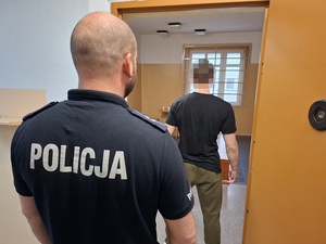 w policyjnej celi stoi zatrzymany mężczyzna. Za nim przy otwartych drzwiach celi stoi policjant służby dyżurnej