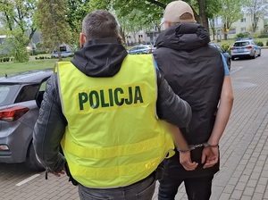 policjant prowadzi w kierunku nieoznakowanego radiowozu mężczyznę, który ma ręce skute za plecami kajdankami