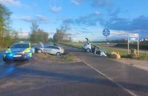 miejsce wypadku drogowego. Na pasie zieleni pomiędzy jezdniami znajduje się ustawiony w poprzek srebrny osobowy samochód z podniesioną maską. Obok na jezdni stoi oznakowany policyjny radiowóz. Po drugiej stronie na poboczu w poprzek jezdni stoi drugi uszkodzony srebrny samochód osobowy