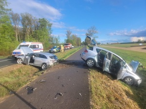 miejsce wypadku drogowego. Na pasie zieleni pomiędzy jezdniami znajduje się ustawiony w poprzek srebrny osobowy samochód z podniesioną maską. Obok na jezdni stoi oznakowany policyjny radiowóz. Po drugiej stronie na poboczu w poprzek jezdni stoi drugi uszkodzony srebrny samochód osobowy. Na jezdni stoi wóz strażacki, karetka i policyjny radiowóz