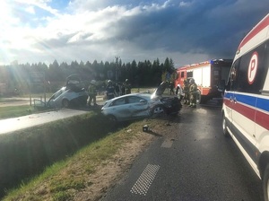 miejsce wypadku drogowego. Na pasie zieleni pomiędzy jezdniami znajduje się ustawiony w poprzek srebrny osobowy samochód z podniesioną maską. Obok na jezdni stoi oznakowany policyjny radiowóz. Po drugiej stronie na poboczu w poprzek jezdni stoi drugi uszkodzony srebrny samochód osobowy. Na jezdni stoi karetka i wóz strażacki. Przy uszkodzonych samochodach stoją strażacy