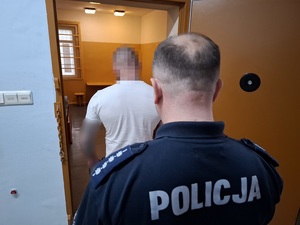 w policyjnej celi stoi zatrzymany mężczyzna. Za nim przy otwartych drzwiach celi stoi policjant służby dyżurnej