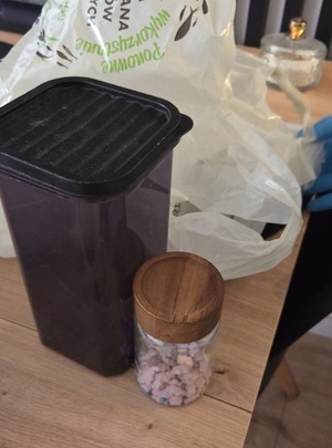 na stole stoi ciemny podłużny plastikowy pojemnik i słój z drewnianą pokrywą wypełniony różowymi tabletkami w kształcie misia