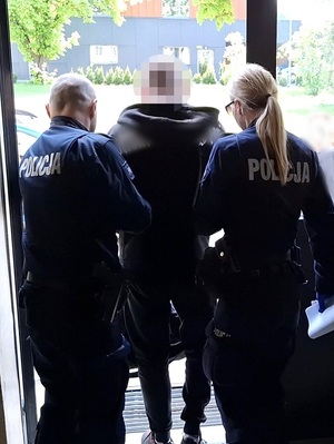 policjant i policjantka służby konwojowej wyprowadzają z budynku komendy zatrzymanego mężczyznę