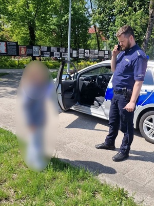 w parku przed dzieckiem i obok radiowozu stoi policjant dzielnicowy, który rozmawia przez telefon komórkowy