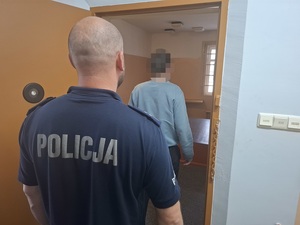 w policyjnej celi stoi zatrzymany mężczyzna. Za nim przy otwartych drzwiach celi stoi policjant służby dyżurnej