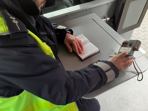 przy stoliku w radiowozie ruchu drogowego siedzi policjant, który trzyma w dłoni alkotest. Na stoliku leży notatnik służbowy