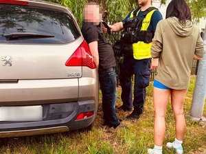 o samochód osobowy opiera się mężczyzna, który ma ręce skute za plecami kajdankami. Policjant poddaje go badaniu alkotestem. Obok stoi kobieta