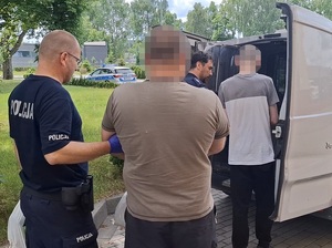 przed otwartymi tylnymi drzwiami nieoznakowanego radiowozu stoją dwaj policjanci oraz dwaj zatrzymani mężczyźni