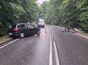 miejsce wypadku drogowego. Na łuku drogi częściowo na jezdni i częściowo na poboczu stoi w poprzek czarny Vw Golf z licznymi uszkodzeniami. Po drugiej stronie, w rowie znajduje się uszkodzony jasny Ford Kuga. Na jezdni stoi policyjny radiowóz
