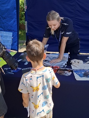 przy policyjnym stoisku profilaktycznym i przy stole z gadżetami stoi policjantka, która rozmawia ze stojącym naprzeciw niej chłopcem