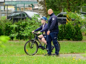 po policyjnym miasteczku ruchu drogowego jedzie rowerem chłopiec. Obok idzie policjant ruchu drogowego