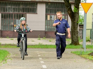 po policyjnym miasteczku ruchu drogowego jedzie rowerem dziewczynka. Obok idzie policjant ruchu drogowego