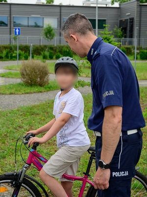 w policyjnym miasteczku ruchu drogowego na oznakowanej drodze znajduje się chłopiec siedzący na rowerze. Przy nim stoi policjant ruchu drogowego