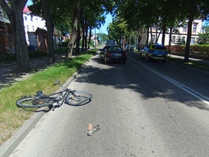 miejsce zdarzenia drogowego. Pora dzienna, słonecznie. Na jezdni stoją obok siebie policyjny radiowóz i czarne osobowe Audi. Za nimi leży uszkodzony rower z wykrzywionymi kołami. Obok na jezdni leży but