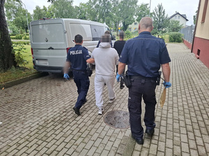 trzej policjanci prowadzą w stronę nieoznakowanego radiowozu służby konwojowej dwóch zatrzymanych mężczyzn