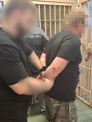 na tle krat policjant kryminalny zakłada kajdanki zatrzymanemu mężczyźnie na ręce trzymane z tyłu