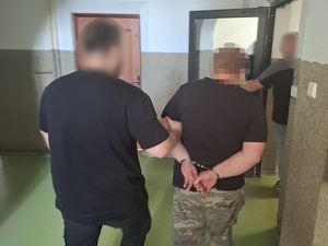 policjant kryminalny prowadzi  mężczyznę, który ma ręce skute za plecami kajdankami w kierunku otwartych drzwi pomieszczenia służbowego