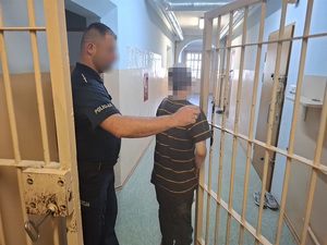 przez okratowane drzwi korytarza pomieszczenia dla osób zatrzymanych przechodzi drobny mężczyzna. Przy drzwiach stoi policjant służby dyżurnej