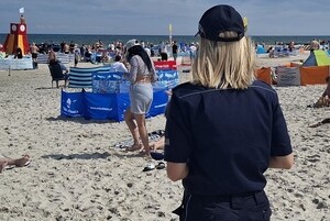 policjantka na nadmorskiej plaży. Na plaży znajdują się parawany i plażowicze. Widać budkę ratowniczą z białą flagą