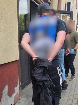dwaj nieumundurowani policjanci wprowadzają do budynku komendy mężczyznę, który ma ręce skute za plecami kajdankami