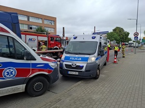 miejsce zdarzenia drogowego. Przy prawej krawędzi jezdni stoi policyjny radiowóz, a przed nim karetka pogotowia. Po drugiej stronie jezdni stoi pojazd ciężarowy z pustą częścią ładunkową. Obok niego stoi wóz straży pożarnej. Pomiędzy autami chodzą policjanci ruchu drogowego i strażacy