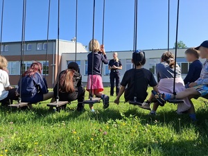 przed grupą dzieci siedzących na placu zabaw stoi policjantka