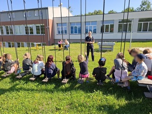 przed grupą dzieci siedzących na placu zabaw stoi policjantka
