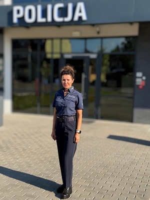 zdjęcie policjantki w letnim umundurowaniu służbowym, stojącej przed budynkiem komisariatu