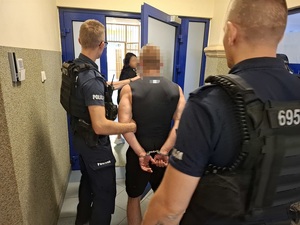 dwaj policjanci służby patrolowej przeprowadzają przez drzwi korytarza pomieszczenia dla osób zatrzymanych mężczyznę, który ma ręce skute za plecami kajdankami