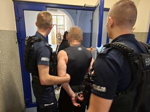 dwaj policjanci służby patrolowej przeprowadzają przez drzwi korytarza pomieszczenia dla osób zatrzymanych mężczyznę, który ma ręce skute za plecami kajdankami