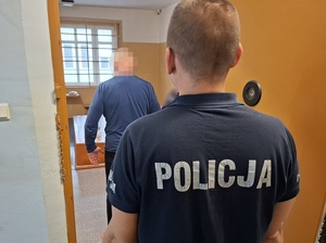 w celi stoi zatrzymany mężczyzna. Za nim, przy otwartych drzwiach celi stoi policjant służby dyżurnej