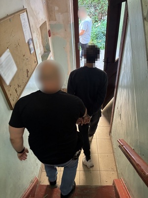 na klatce schodowej policjant kryminalny sprowadza schodami w kierunku wyjścia mężczyznę, który ma ręce skute za plecami kajdankami