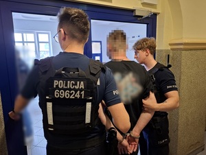 przed drzwiami wyjściowymi prowadzącymi do poczekalni komendy stoją dwaj policjanci oraz mężczyzna, który ma ręce skute za plecami kajdankami