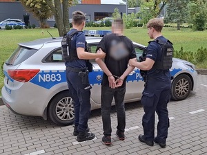 przed oznakowanym radiowozem stoją dwaj policjanci przytrzymujący mężczyznę, który ma ręce skute za plecami kajdankami