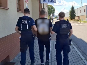 w stronę wejścia do budynku komendy Policji idą dwaj policjanci służby patrolowej, prowadzący mężczyznę, który ma ręce skute za plecami kajdankami