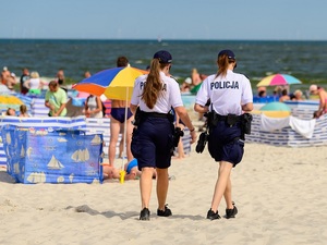 nadmorską plażą wśród plażowiczów idą dwie policjantki ogniwa wodnego ubrane w białe koszulki polo i czarne krótkie spodenki
