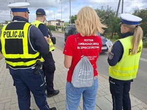 przed przejazdem kolejowym stoi dwoje policjantów ruchu drogowego. Między nim stoi przedstawicielka kolei w czerwonej koszulce z białym napisem na plecach "Jestem ambasadorem bezpieczeństwa"