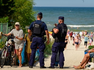 przed wejściem na plażę nad morzem stoją dwaj policjanci. Obok znajdują się przechodnie i plażowicze