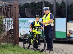 przy bramie na tle autokaru stoi policjantka ruchu drogowego, a obok niej chłopiec z rowerem, z kaskiem na głowie i w kamizelce odblaskowej