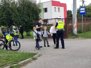 przed bramą prowadzącą na teren szkoły i obok przystanku autobusowego policjantka ruchu drogowego rozmawia z młodym mężczyzną, starszą kobietą i chłopcem. W stronę szkoły idą uczniowie
