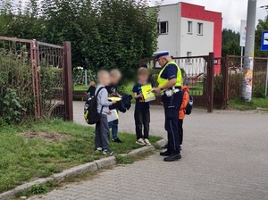 przed bramą szkoły policjantka ruchu drogowego rozdaje kamizelki odblaskowe trzem chłopcom
