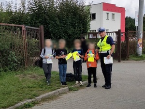 przed bramą szkoły stoi policjantka ruchu drogowego, a obok niej czterej chłopcy trzymający opakowania z kamizelkami odblaskowymi