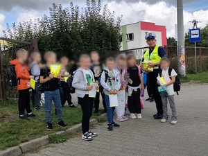 przed bramą szkoły, w towarzystwie grupy uczniów młodszych klas stoi policjantka ruchu drogowego. Dzieci trzymają kamizelki odblaskowe