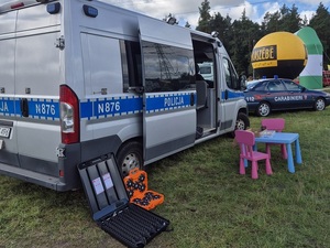 na placu na trawie stoi oznakowany policyjny radiowóz typu bus. Obok otwartych drzwi leżą: skrzynka z kolczatką drogową i skrzynka z flarami drogowymi. Przed radiowozem stoi dziecięcy stolik i krzesełka, na stoliku materiały pliśmiennicze